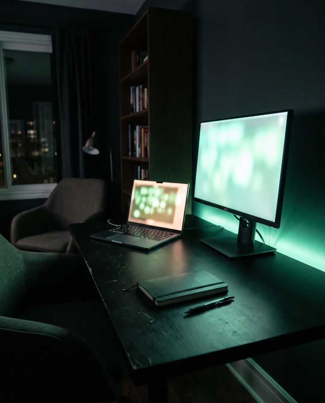 Escritorio oscuro de noche con una notebook abierta y un monitor encendidos, los dos fuera de foco y con luz verde, un cuaderno cerrado y una birome al lado