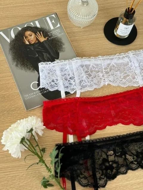 Tres portaligas de encaje apoyados uno al lado del otro —blanco, rojo y negro—, cada uno con sus ligas y sus broches, sobre una mesa de madera clara.