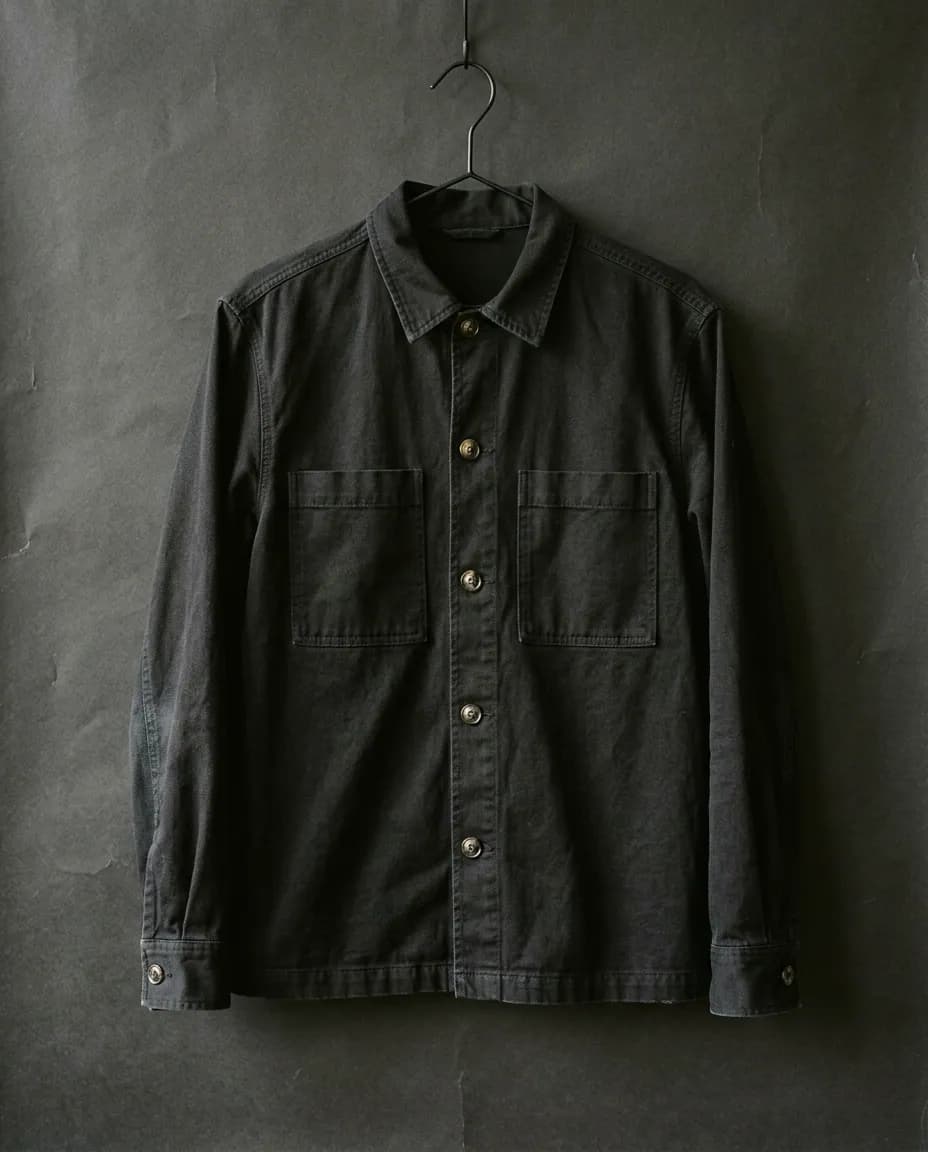 Camisa de gabardina negra colgada de una percha delgada contra un fondo de papel oscuro