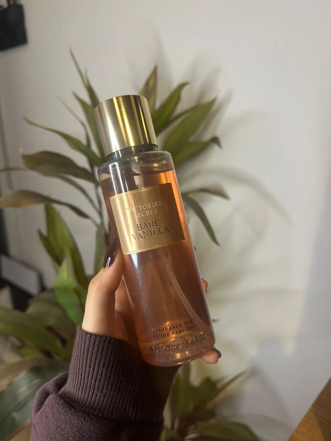 Frasco de body splash Victoria’s Secret Bare Vanilla, dorado claro, sostenido en la mano frente a una planta