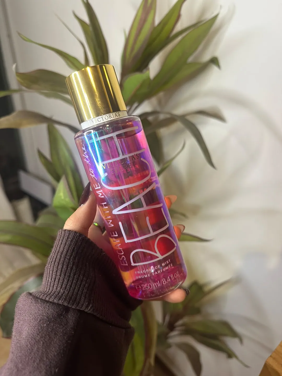 Frasco de body splash Victoria’s Secret Beach, tornasolado rosa y violeta, sostenido en la mano frente a una planta