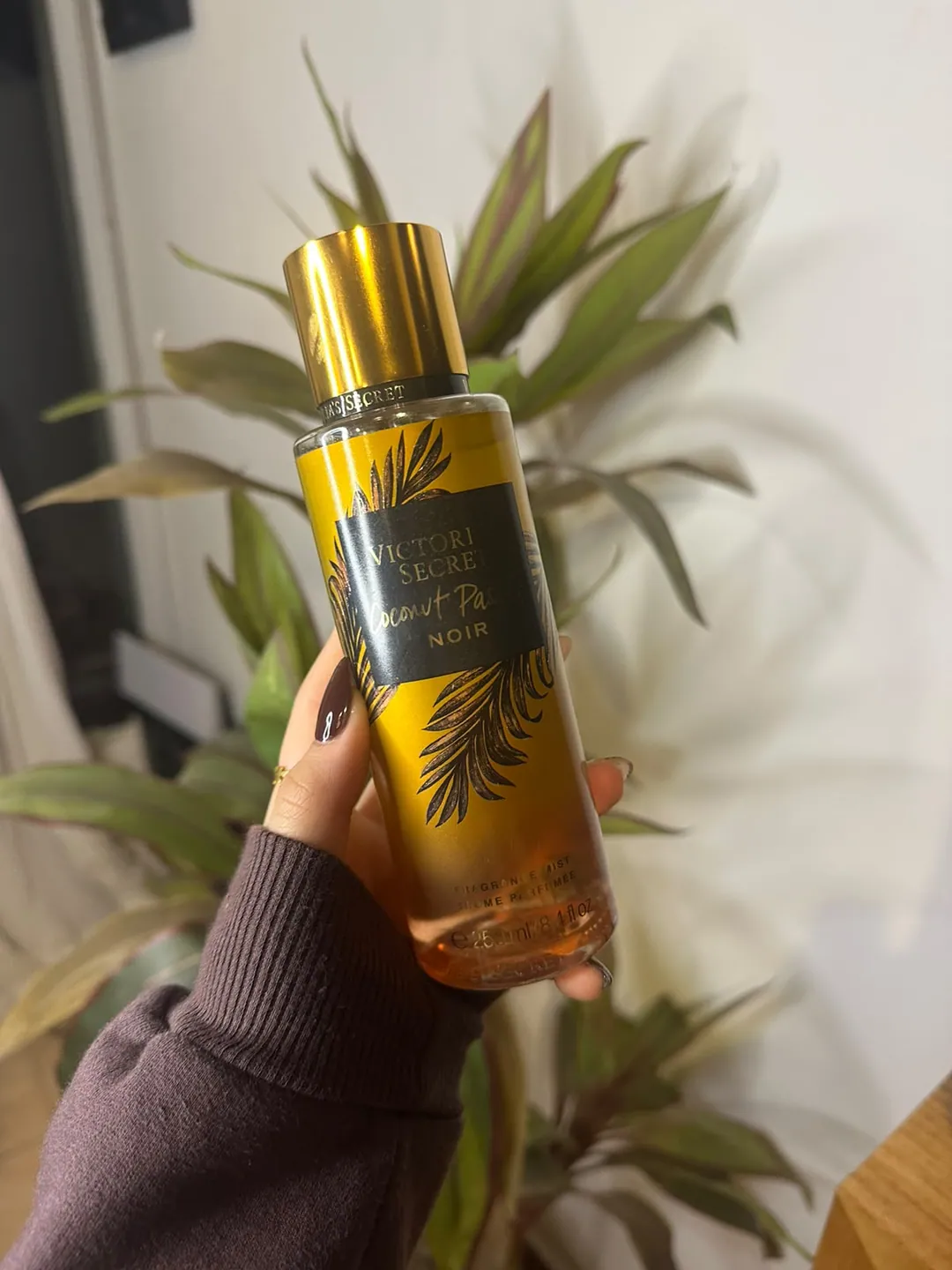 Frasco de body splash Victoria’s Secret Coconut Passion Noir, dorado con hojas de palmera y etiqueta negra, sostenido en la mano