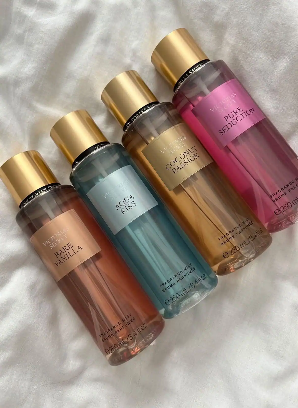 Cuatro brumas de Victoria’s Secret —Bare Vanilla, Aqua Kiss, Coconut Passion y Pure Seduction— apoyadas en diagonal sobre una tela blanca