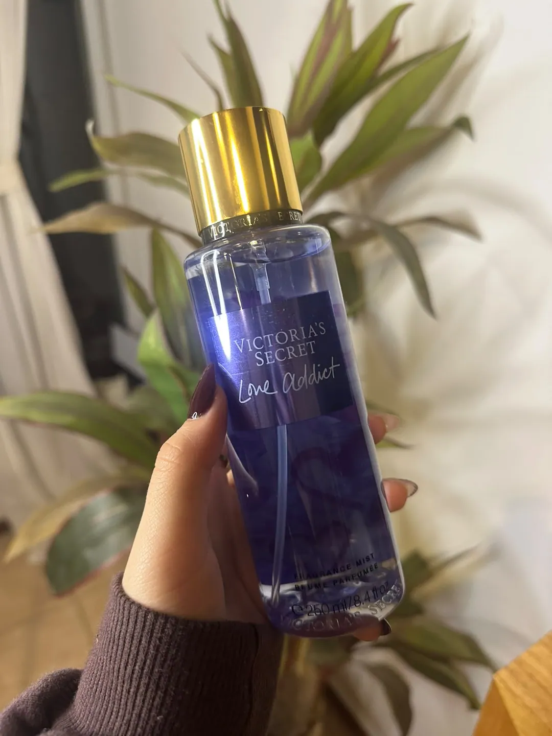 Frasco de body splash Victoria’s Secret Love Addict, azul violáceo, sostenido en la mano frente a una planta