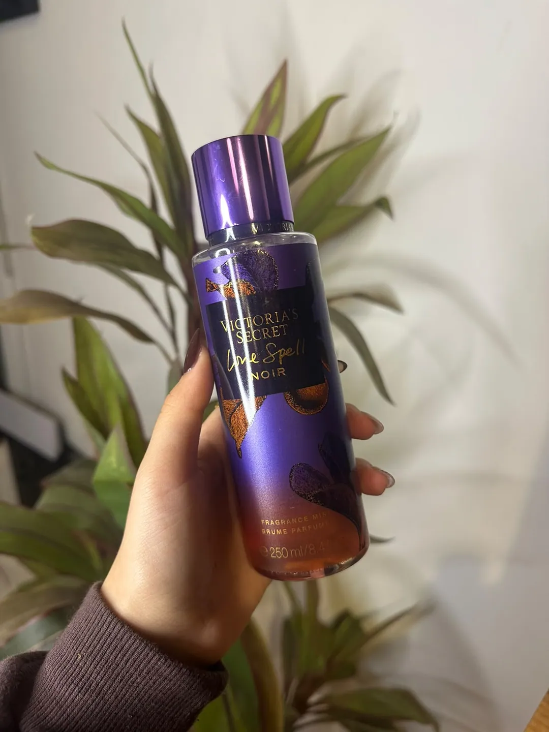 Frasco de body splash Victoria’s Secret Love Spell Noir, violeta con hojas doradas, sostenido en la mano