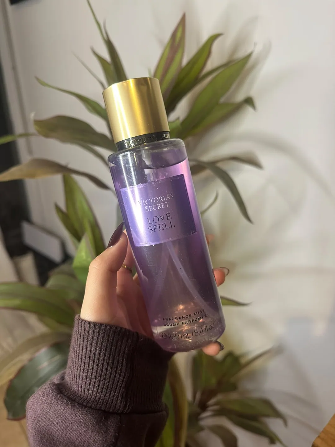 Frasco de body splash Victoria’s Secret Love Spell, lila, sostenido en la mano frente a una planta