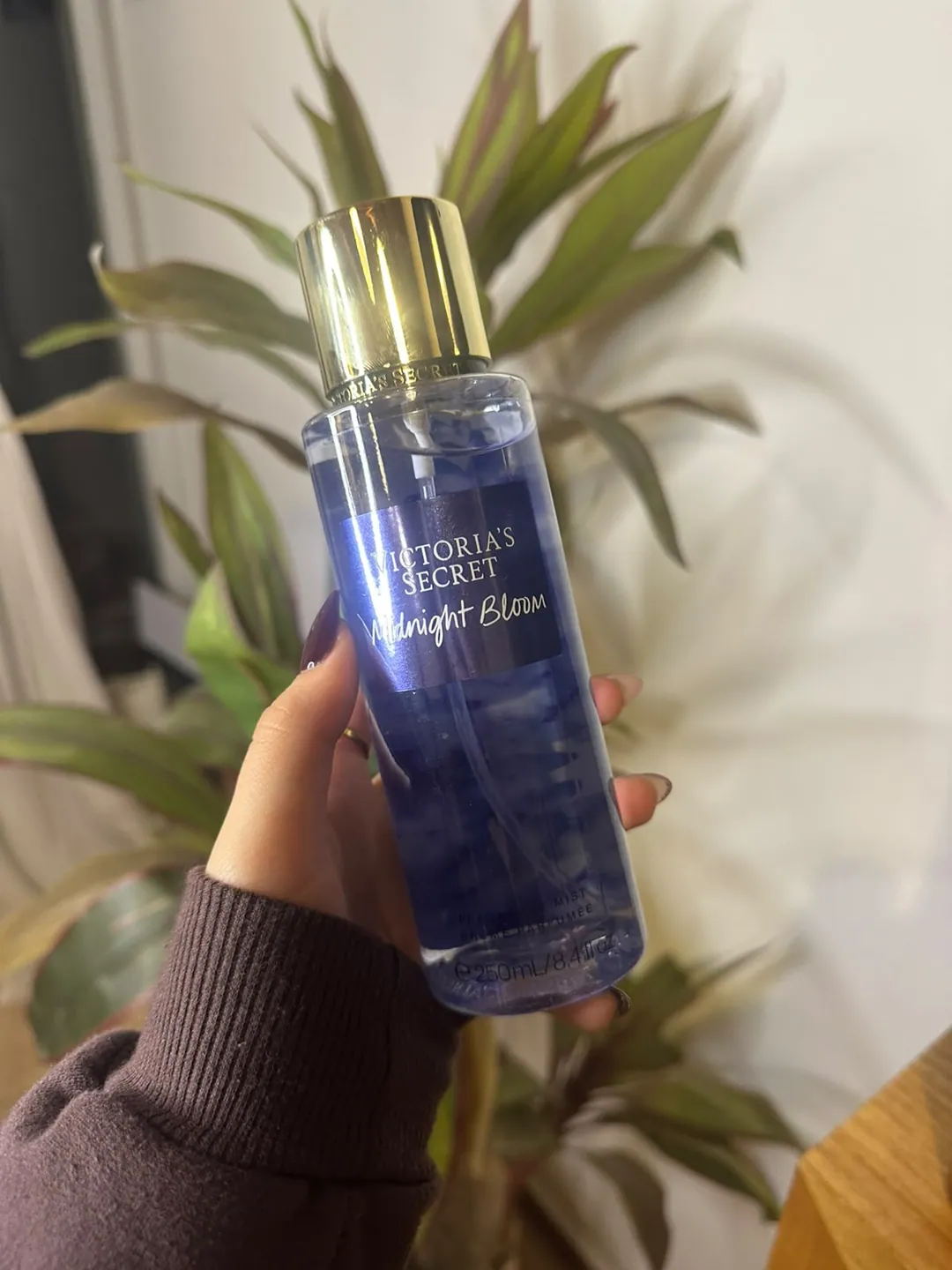 Frasco de body splash Victoria’s Secret Midnight Bloom, azul noche, sostenido en la mano frente a una planta