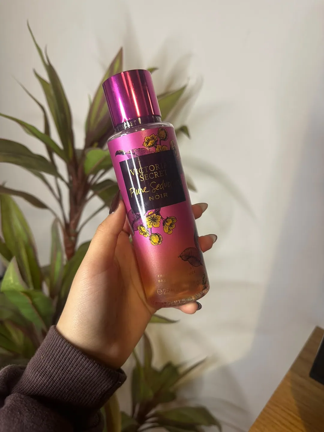 Frasco de body splash Victoria’s Secret Pure Seduction Noir, fucsia con flores doradas y etiqueta negra, sostenido en la mano