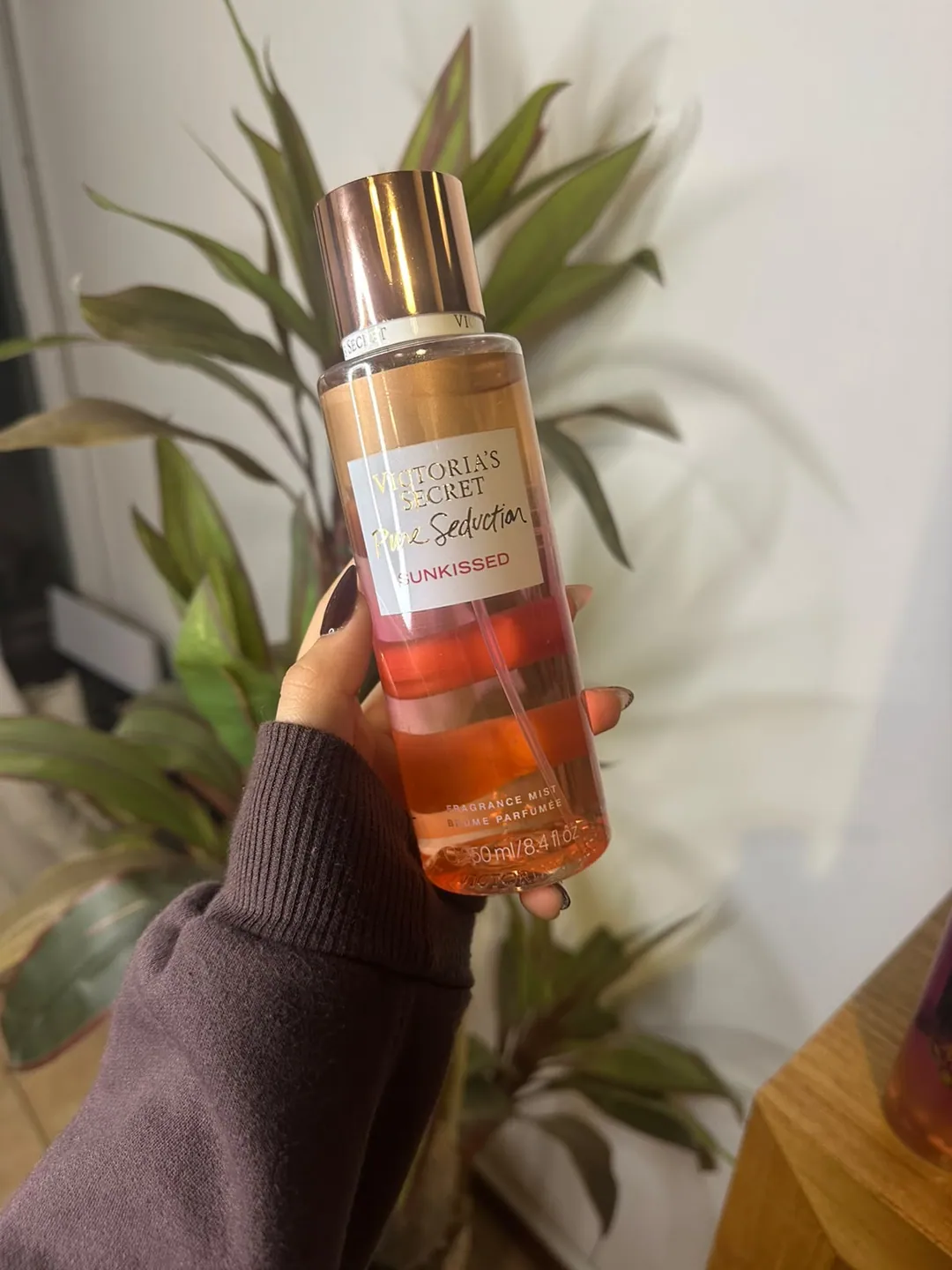 Frasco de body splash Victoria’s Secret Pure Seduction Sunkissed, degradé durazno y ámbar, sostenido en la mano frente a una planta