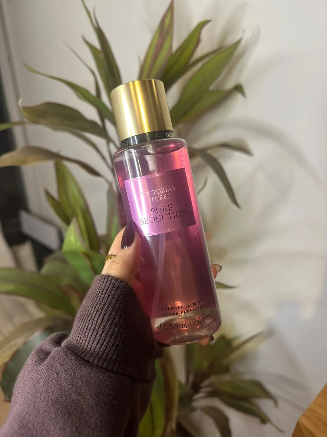 Frasco de body splash Victoria’s Secret Pure Seduction, rosa intenso, sostenido en la mano frente a una planta
