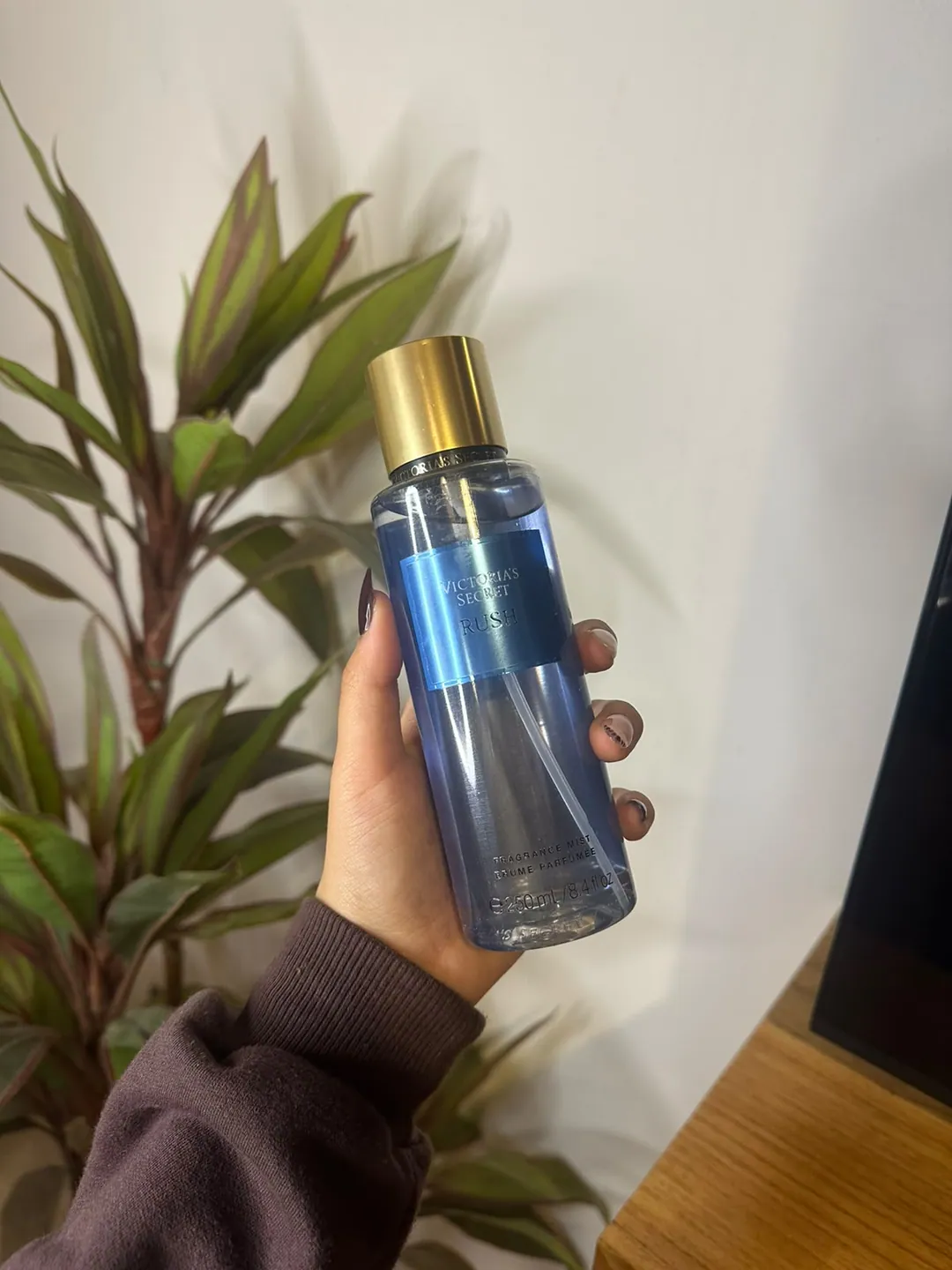 Frasco de body splash Victoria’s Secret Rush, azul, sostenido en la mano frente a una planta